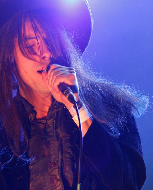 Aldous RH, un homme au longs cheveux noirs avec un chapeau noir sur la scène du Patchwork Festival